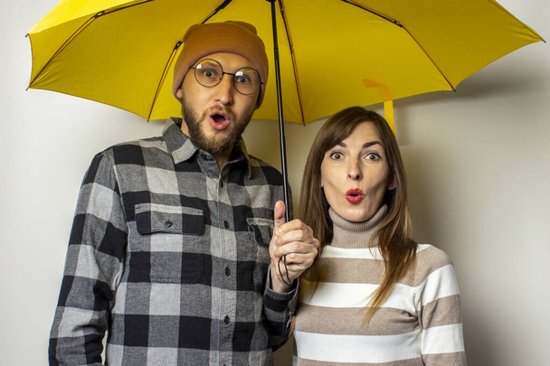 Ein junges Paar mit überraschten Gesichtern, ein Mann mit Bart, Hut und kariertem Hemd und ein Mädchen im Pullover unter einem gelben Regenschirm auf einem isolierten hellen Hintergrund. Emotionales Gesicht. Glückliches Paar. - Foto, Bild