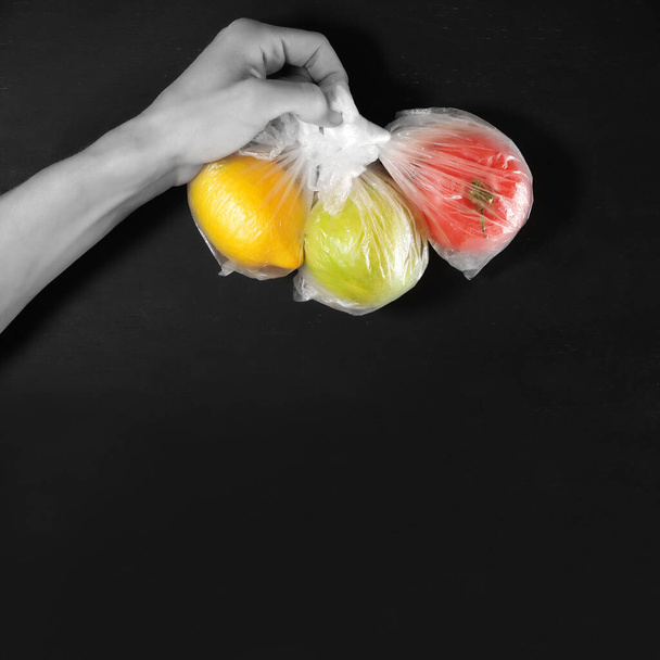 hand holds fruits apple, tomato and lemon, each in individual plastic package, environment pollution problem in quarantine isolation, concept of safety delivery, black background with copy space - Photo, Image