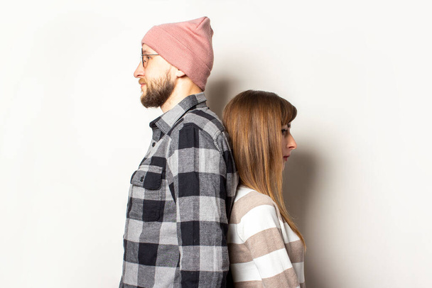 Ένας νεαρός με γενειάδα με καπέλο και καρό πουκάμισο και ένα κορίτσι με πουλόβερ στέκονται πλάτη με πλάτη σε ένα απομονωμένο φόντο φωτός. Συναισθηματικό πρόσωπο. - Φωτογραφία, εικόνα