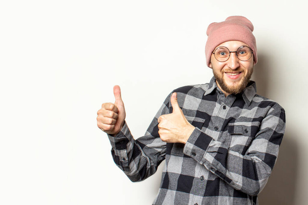 Portrait of a young man with a beard in a hat, a checkered shirt makes a gesture thumb up on an isolated light background. Emotional face. The gesture is all right, OK, verified. - Photo, Image