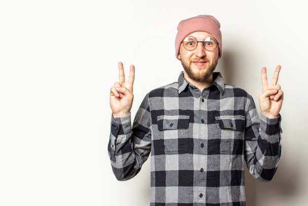 Portrait of a young man with a beard in a hat, a checkered shirt makes a gesture two fingers up on an isolated light background. Emotional face. Gesture greeting, Peace. - Фото, изображение