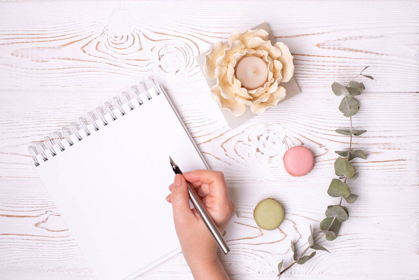 Uma mão feminina segura uma caneta-tinteiro sobre um diário limpo com primavera, macaroons e um castiçal em forma de flor em uma mesa de madeira rasgada branca. Moda flat lay, lugar para texto
 - Foto, Imagem