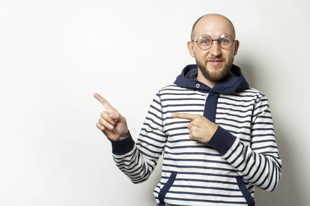 Portrait of a Bald Young man with a beard in glasses of a jacket with a hood points a finger to the side on an isolated light background. Emotional face. Gesture of shock, surprise. - Photo, Image