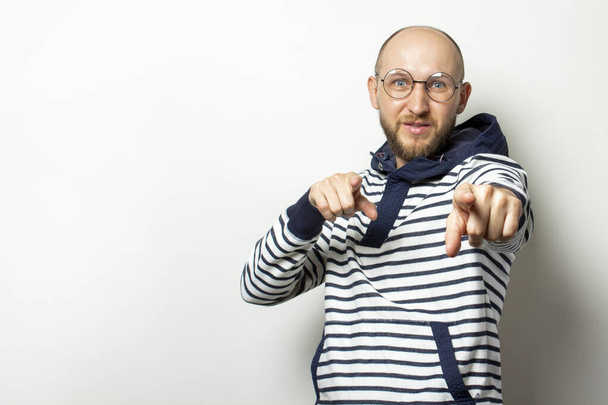Portrait of a Bald Young man with a beard in glasses, a sweater with a hood, points his fingers at the viewer on an isolated light background. Emotional face. - Photo, Image
