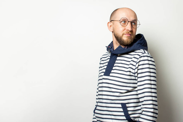 Portrait of a Bald Young man with a beard in glasses, a sweater with a hood looks back on an isolated light background. Emotional face. Gesture looking back. - Photo, Image