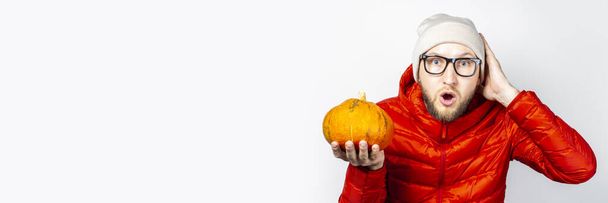 испуганный молодой человек в красной куртке, шляпе, держит тыкву и держит руку к голове на светлом фоне. Концепция Хэллоуина, праздника, осени. Баннер. - Фото, изображение