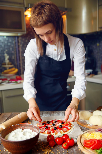 Woman in white shirt and apron putting the ingredients on the pizza. Pizza concept. Production and delivery of food - Zdjęcie, obraz