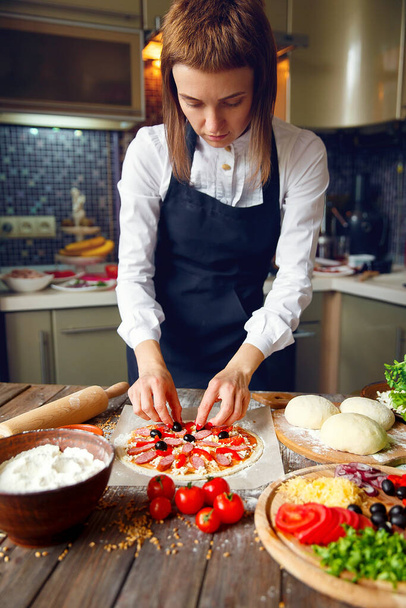 Woman in white shirt and apron putting the ingredients on the pizza. Pizza concept. Production and delivery of food - Foto, Bild