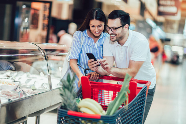 Ευτυχισμένο ζευγάρι νέων δεσμών μεταξύ τους και χαμογελώντας, ενώ το περπάτημα, ενώ το περπάτημα στο κατάστημα τροφίμων με καλάθι αγορών χρησιμοποιώντας έξυπνο τηλέφωνο - Φωτογραφία, εικόνα