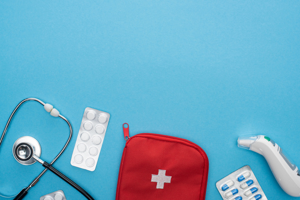 bovenaanzicht van pillen in blisterverpakkingen, stethoscoop, verbanddoos en oorthermometer op blauwe achtergrond - Foto, afbeelding