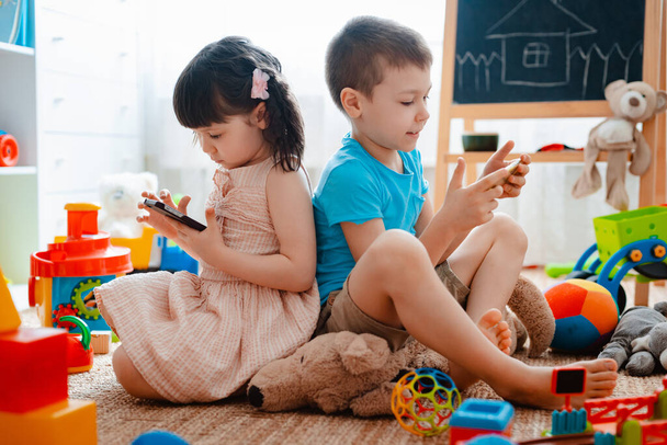 Sourozenci děti bratr a sestra, přátelé sedí na podlaze domu v dětském pokoji s chytrými telefony, odděleny od roztroušených hraček. - Fotografie, Obrázek