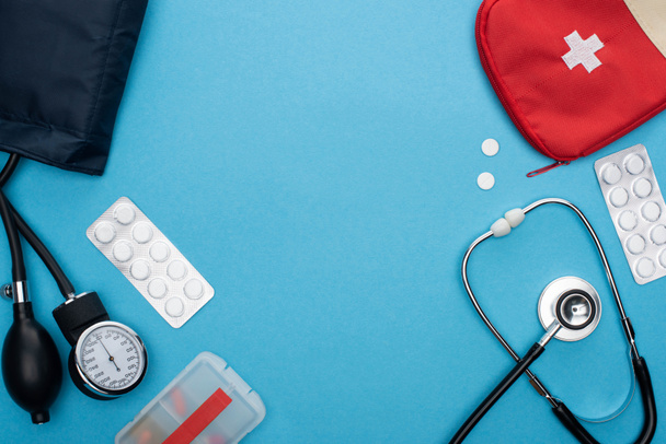 bovenaanzicht van pillen in blisterverpakkingen, sfygmomanometer, verbanddoos en stethoscoop op blauwe achtergrond - Foto, afbeelding