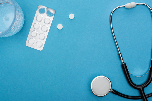 Ansicht von Pillen in Blisterverpackung, Wasserglas und Stethoskop auf blauem Hintergrund - Foto, Bild