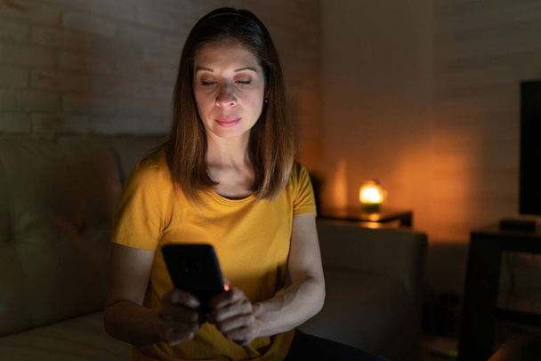 Dobře vypadající žena ve 40s pomocí smartphonu textovat a kontrolovat zprávy, zatímco sedí v obývacím pokoji v noci - Fotografie, Obrázek