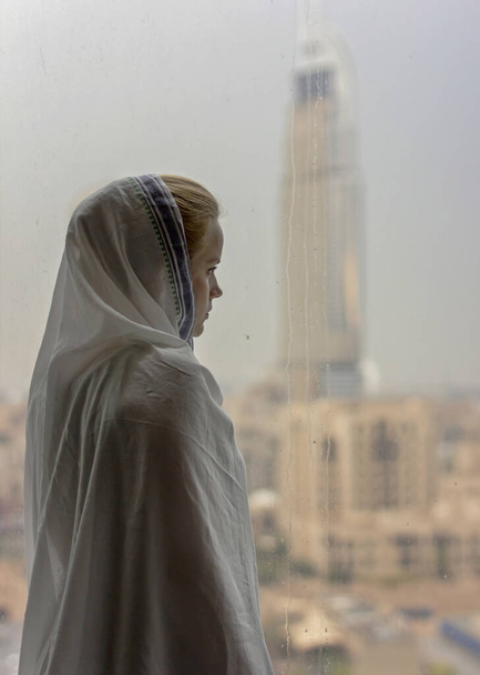Una joven en un hiyab mira por la ventana. Una mujer en un país musulmán. Una chica con la cabeza cubierta. Figura femenina junto a la ventana. Una mujer está mirando la ciudad
. - Foto, imagen
