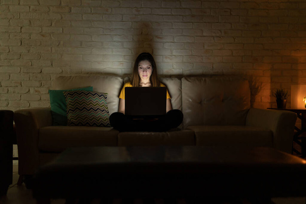 Breed zicht op een vrouw die in een bank zit en 's nachts vanuit huis werkt met een laptop - Foto, afbeelding