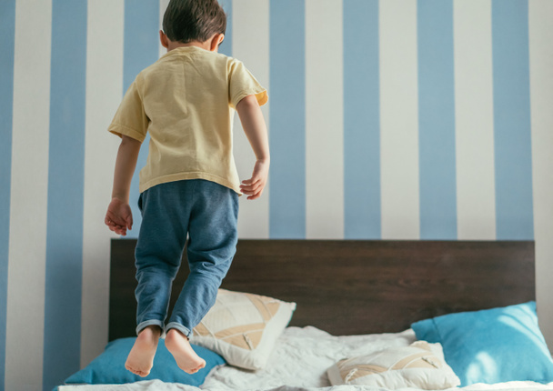 back view of barefoot boy in blue jeans jumping on bed at home - Fotografie, Obrázek
