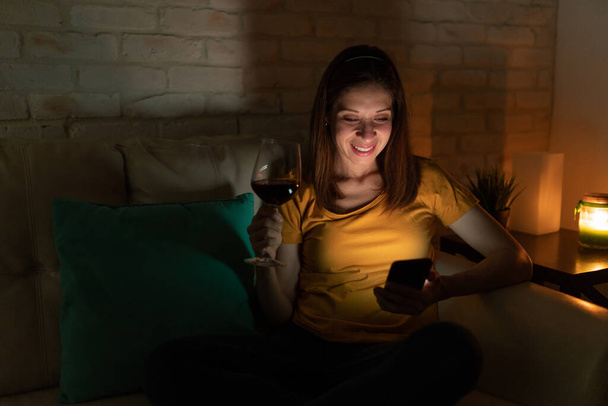Portret szczęśliwej brunetki smsującej ze swoim partnerem na smartfonie podczas nocnego picia lampki wina - Zdjęcie, obraz