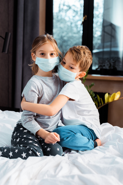 χαριτωμένος αδελφός αγκαλιάζει αδελφή σε ιατρική μάσκα, ενώ κάθεται στο κρεβάτι - Φωτογραφία, εικόνα