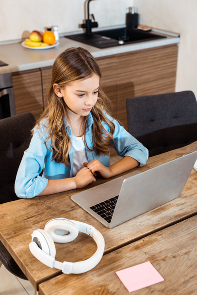 Nettes Kind lernt online, während es zu Hause Laptop in der Nähe von Kopfhörern anschaut - Foto, Bild