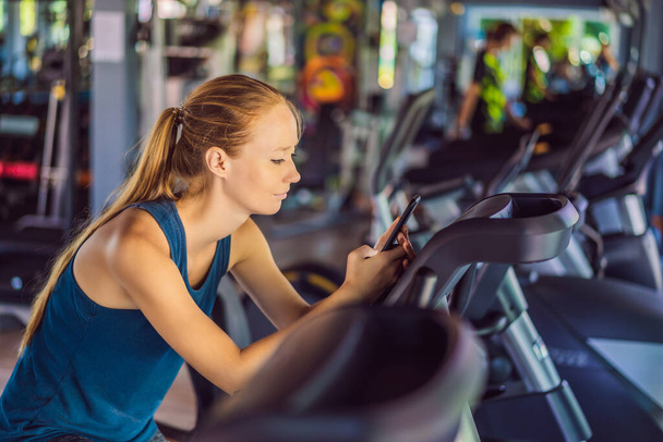 Coronavirus is over. Quarantine weakened. Take off the mask. Now you can go to public places. Young woman using phone while training at the gym. Woman sitting on exercising machine holding mobile - Foto, Imagem