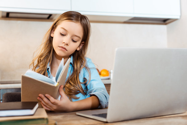 valikoiva painopiste söpö lapsi tilalla kirja lähellä kannettavaa tietokonetta opiskellessaan verkossa kotona
 - Valokuva, kuva