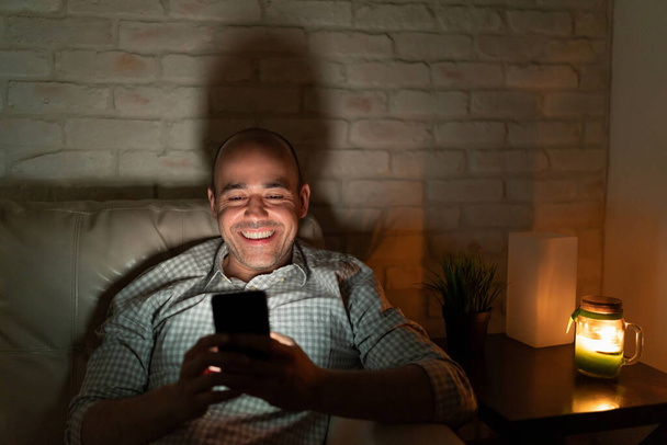 Chico feliz enviando mensajes a su novia mientras se relaja en casa por la noche y sonríe
 - Foto, Imagen