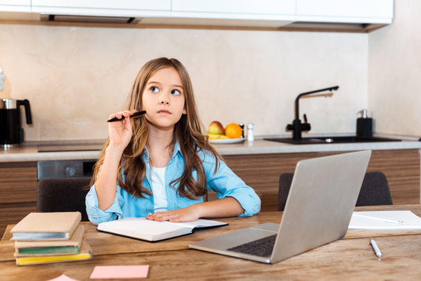 Evde e-öğrenim görürken dizüstü bilgisayarın yanında kalem tutan düşünceli çocuğun seçici odağı - Fotoğraf, Görsel