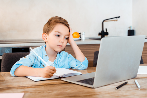 επιλεκτική εστίαση βαριέται αγόρι γραπτώς στο σημειωματάριο, ενώ κοιτάζοντας φορητό υπολογιστή και e-learning στο σπίτι - Φωτογραφία, εικόνα