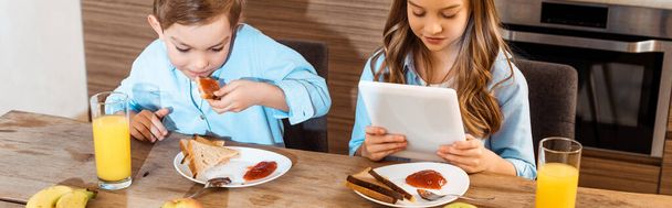 デジタルタブレットを使って妹のそばでジャムパンを食べる男の子のパノラマ作物  - 写真・画像