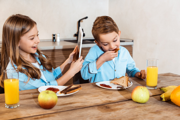 happy kid using digital tablet near brother eating toast bread with jam  - 写真・画像