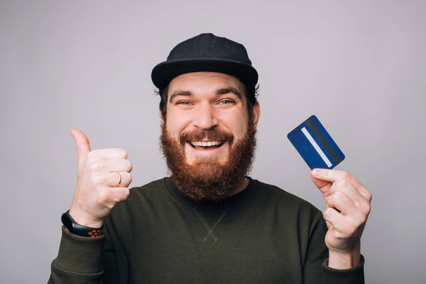 Zadowolony brodaty mężczyzna pokazuje kciuk w górę i niebieską kartę kredytową lub debetową. - Zdjęcie, obraz