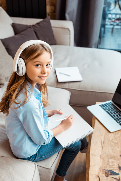 glückliches Kind in drahtlosen Kopfhörern, das in einem Notizbuch neben dem Laptop mit leerem Bildschirm schreibt, während es zu Hause E-Learning macht - Foto, Bild