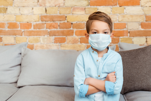 chlapec v lékařské masce se zkříženými pažemi v blízkosti cihlové zdi v obývacím pokoji  - Fotografie, Obrázek