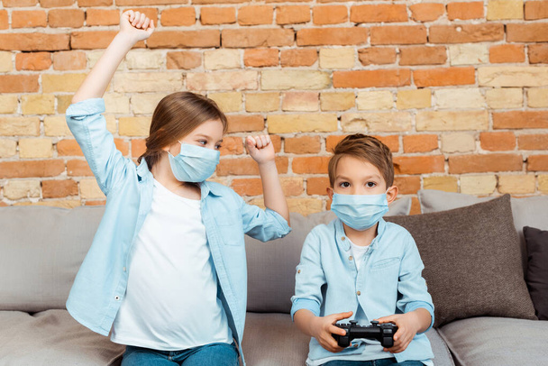 KYIV, UKRAINE - APRIL 27, 2020: sister gesturing near brother in medical mask playing video game - Photo, Image