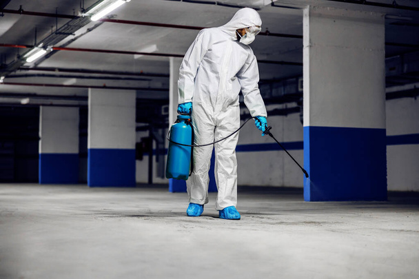 Pełna długość pracownika w sterylnym kombinezonie ochronnym i masce dezynfekującej podziemny garaż przed wirusem korony / covid-19. - Zdjęcie, obraz