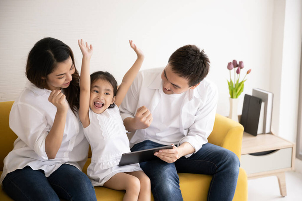 Ευτυχισμένη ασιατική οικογένεια που χρησιμοποιεί τεχνολογίες για διασκέδαση. Πατέρας, μητέρα και κόρη κοιτάζουν ψηφιακή πλακέτα στον καναπέ του σπιτιού. Τεχνολογία επικοινωνίας και τρόπος ζωής - Φωτογραφία, εικόνα