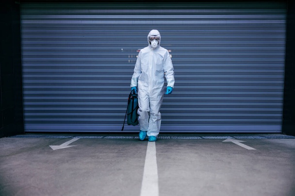 Mann in sterilem Schutzanzug und Maske hält Desinfektionsmittel in der Hand und geht in Tiefgarage auf Kamera zu. - Foto, Bild