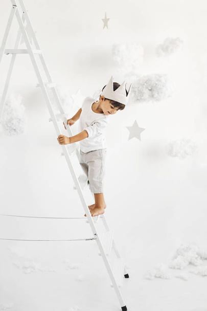 portrait of a cute little boy in crown on white ladder among clouds on a white background - Foto, Imagem