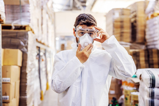 Молода людина в захисному костюмі кладе захисні окуляри і готується до дезінфекції від коронавірусу / ковад-19. Складський інтер'єр. Склад наповнений продуктами харчування
. - Фото, зображення