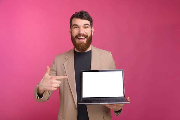 Excited bearded man is pointing at his laptop he is holding on pink background. - Фото, изображение
