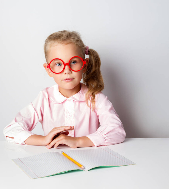 Klein blond meisje met bril en roze blouse. Zittend aan tafel met kopieerboek en potlood erop. Poseren geïsoleerd op wit - Foto, afbeelding