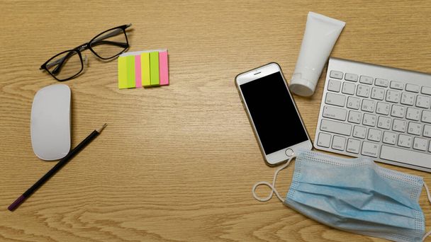 Horní pohled na bílou počítačovou klávesnici, myš, tužku, smartphone, alkoholový gel, masku, brýle a barevný papírový pohled na pracovní stůl z hnědého dřeva. pro práci z domova nebo WFH - Fotografie, Obrázek