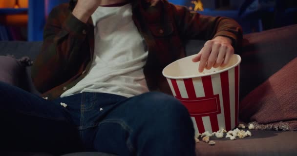 Crop view of male person eating popcorn from paper bowl while sitting on sofa and watching tv. Concept of leisure and entertainment. Mess on couch. - Filmagem, Vídeo