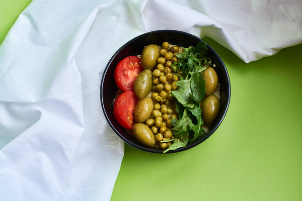 Green summer background and white cloth with  black bowl and delicious useful breakfast of porridge and vegetables. Tomatoes, olives, arugula, peas. - Photo, Image
