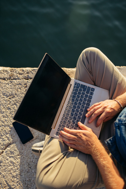 Wolny strzelec korzystający z laptopa na plaży. Mężczyzna siedzący na plaży i pracujący zdalnie przy użyciu laptopa i łącza internetowego. - Zdjęcie, obraz