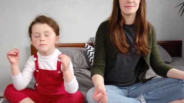 Healthy calm young mother teaching meditation sit with cute small child daughter on bed at home, cheerful happy mom and kid girl doing yoga exercises relaxing together in lotus pose - Video
