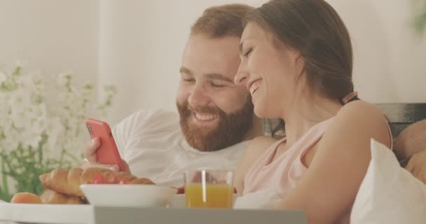 Close up view of couple looking at smartphone screen and smiling while spending time together in bed. Happy girl and man scrolling social media news feed while having breakfast. - Кадры, видео