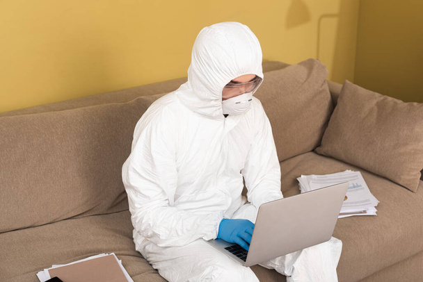 Man in medical mask, hazmat suit and safety goggles using laptop near documents on couch  - Photo, Image