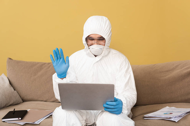 Hombre en traje de materiales peligrosos y máscara médica que tiene videollamada en el ordenador portátil cerca de documentos en el sofá
  - Foto, imagen